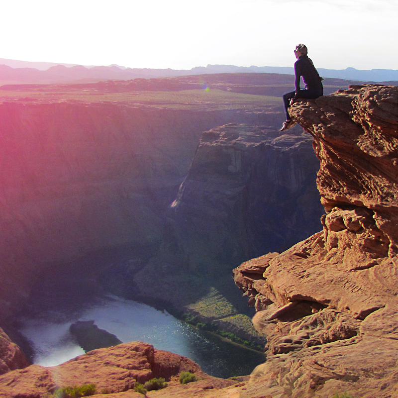 Taylor George sitting on the edge of a red rock cliff overlooking the river and valley below